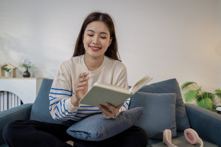 A young woman is sitting comfortably on the sofa, smiling while reading a book with focus. Her...