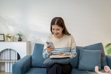 A young woman sitting comfortably on a sofa holding a smartphone while smiling and reading a...