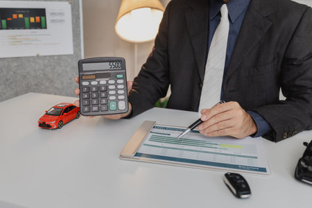 A businessman in a suit is calculating car loan options using a calculator and comparison chart, with model cars and car keys on the desk, symbolizing finance, leasing, and investment planning.の写真素材