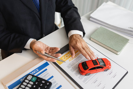 A businessman in a formal suit explains car insurance details, holding a small red car model and pointing at contract documents, symbolizing agreement, protection, and financial planning.の写真素材