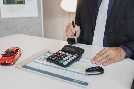 A businessman in a suit is calculating car loan options using a calculator and comparison chart, with model cars and car keys on the desk, symbolizing finance, leasing, and investment planning.の写真素材