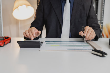 A businessman in a suit is calculating car loan options using a calculator and comparison chart, with model cars and car keys on the desk, symbolizing finance, leasing, and investment planning.の写真素材