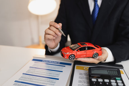 A businessman in a formal suit explains car insurance details, holding a small red car model and pointing at contract documents, symbolizing agreement, protection, and financial planning.の写真素材