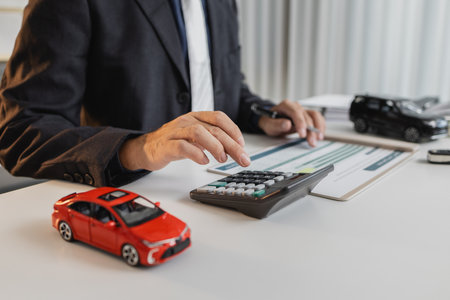 A businessman in a suit is calculating car loan options using a calculator and comparison chart, with model cars and car keys on the desk, symbolizing finance, leasing, and investment planning.の写真素材