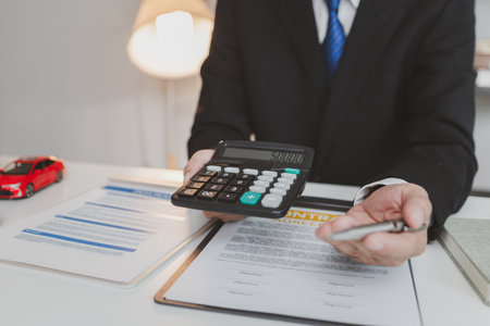 A businessman in a formal suit explains car insurance details, holding a small red car model and pointing at contract documents, symbolizing agreement, protection, and financial planning.の写真素材