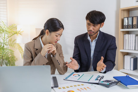 Two business professionals are discussing financial charts and reports in a modern office. They collaborate, share ideas, and analyze data together, highlighting teamwork and strategy.の写真素材
