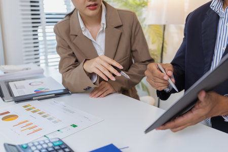 Two business professionals are discussing financial charts and reports in a modern office. They collaborate, share ideas, and analyze data together, highlighting teamwork and strategy.の写真素材
