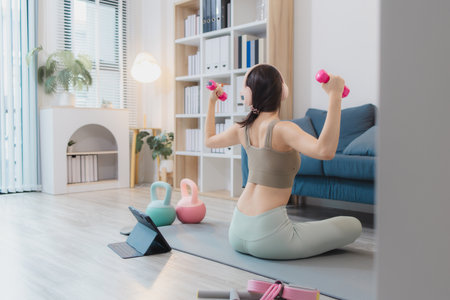 A young woman is exercising at home, practicing yoga and strength training with dumbbells and kettlebells on a mat. She follows her workout routine with focus and determination.の写真素材