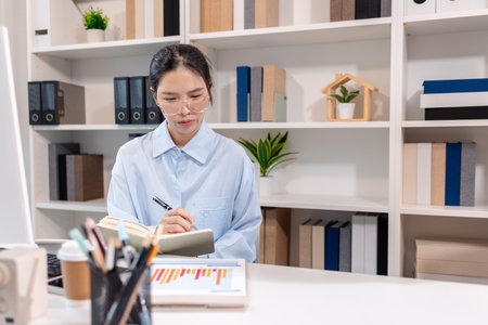 A young Asian businesswoman is sitting at her desk in a modern office, reviewing printed documents while working on a computer, showing focus, dedication, and professional attitude.の写真素材