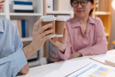 Two Asian women in a startup office clinking coffee cups to celebrate teamwork and successful project collaboration, symbolizing partnership, friendship, and positive workplace culture.の写真素材
