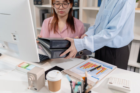 Two female office employees are discussing tasks while one of them hands over two large binders, symbolizing workload delegation and teamwork in a modern corporate environment.の写真素材