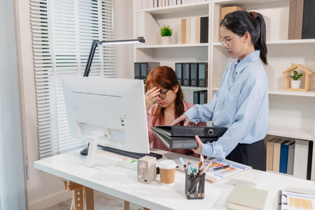 Two female colleagues working in an office, one looking stressed with her hand on her forehead while the other offers a folder, symbolizing teamwork and workplace pressure in a busy department.の写真素材