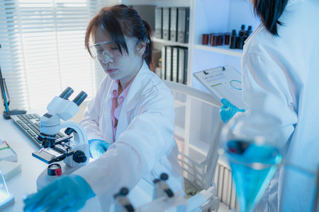 Two female scientists in white lab coats working together in a bright laboratory with microscopes, glassware, and blue test tubes, representing teamwork and modern scientific research environment.の写真素材