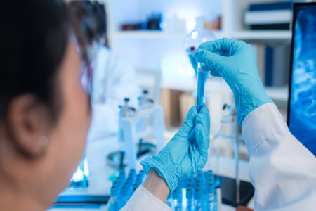 Two female scientists in white lab coats working together in a bright laboratory with microscopes, glassware, and blue test tubes, representing teamwork and modern scientific research environment.の写真素材