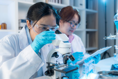 Two female scientists in white lab coats working together in a bright laboratory with microscopes, glassware, and blue test tubes, representing teamwork and modern scientific research environment.の写真素材