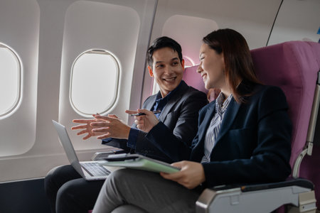Two Asian business professionals smiling and discussing ideas during a flight, sitting next to each other in an airplane cabin. The man gestures confidently while the woman listens attentively.の写真素材