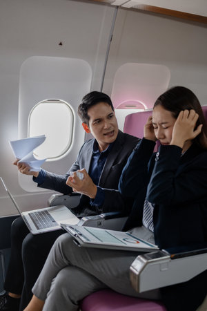 Two Asian business professionals in a heated discussion during a flight. man points at documents with frustration, while the woman covers her ears in stress, sitting in airplane seats with a laptop.の写真素材
