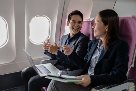 Two Asian business professionals smiling and discussing ideas during a flight, sitting next to each other in an airplane cabin. The man gestures confidently while the woman listens attentively.の写真素材