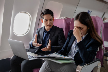 Frustrated Asian business colleagues on a plane facing a stressful situation. man is explaining something with a serious expression while the woman holds her face in distress, overwhelmed by tension.の写真素材
