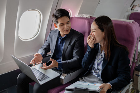 Two Asian business professionals in formal suits discussing work mid-flight. Seated on an airplane, they are exchanging ideas with expressive hand gestures and attentive facial expressions.の写真素材