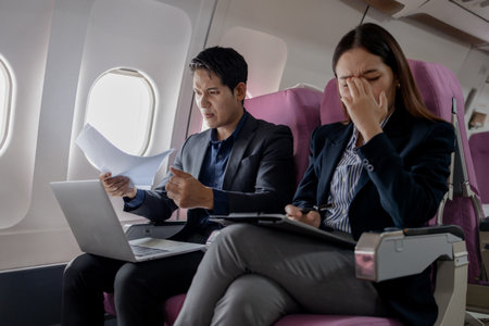 Two Asian business professionals in a heated discussion during a flight. man points at documents with frustration, while the woman covers her ears in stress, sitting in airplane seats with a laptop.の写真素材