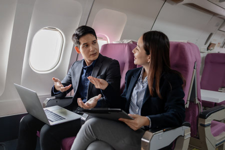 Two Asian business professionals in formal suits discussing work mid-flight. Seated on an airplane, they are exchanging ideas with expressive hand gestures and attentive facial expressions.の写真素材