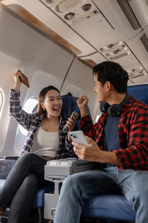 Excited young Asian couple sitting on an airplane, holding a passport, cash, and smartphone while celebrating together. The woman looks happy, and the man shows a cheering gesture, enjoying the momentの写真素材