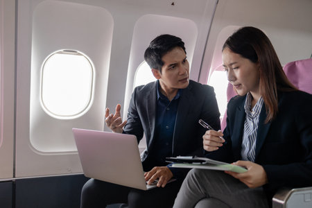 Two Asian business professionals discussing work during a flight, sitting in an airplane cabin. The man gestures seriously while the woman listens and holds documents and a pen, appearing focused.の写真素材