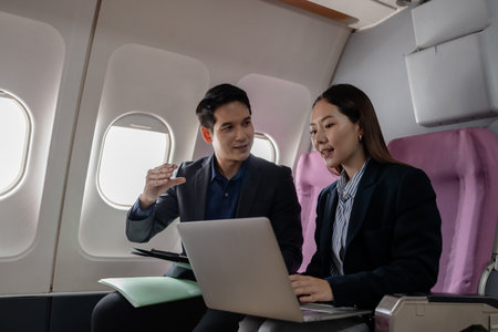 Asian business professionals working together on a laptop while sitting on a commercial airplane. They appear to be discussing a project or presentation in a productive and collaborative manner.の写真素材