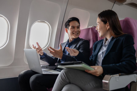Two Asian business professionals smiling and discussing ideas during a flight, sitting next to each other in an airplane cabin. The man gestures confidently while the woman listens attentively.の写真素材