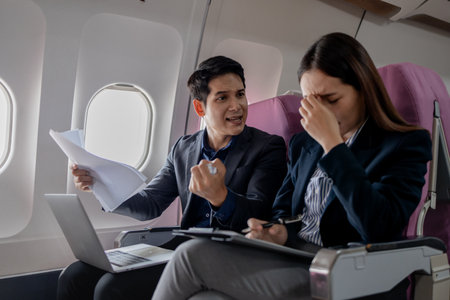 Two Asian business professionals in a heated discussion during a flight. man points at documents with frustration, while the woman covers her ears in stress, sitting in airplane seats with a laptop.の写真素材