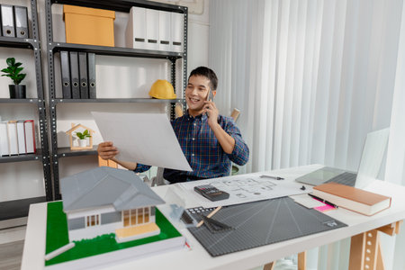 Confident young architect working at office desk, drawing house plans and discussing construction details on the phone, focusing on accuracy and creativity in architectural design and project planningの写真素材