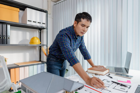 Young architect working in office with building models and floor plans, analyzing details and...