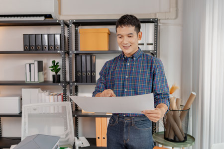 A young architect works with building plans and a house model in the office, reviewing blueprints, marking details, and analyzing design structures with focus and creativity.の写真素材