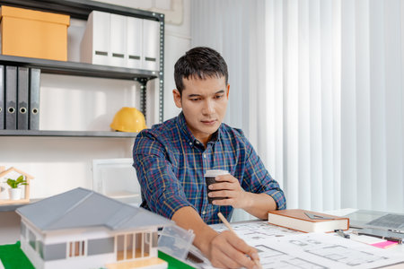 A young architect is working at his desk, reviewing blueprints, using a calculator, and checking a house model. He focuses on precision and detail while planning construction projects.の写真素材