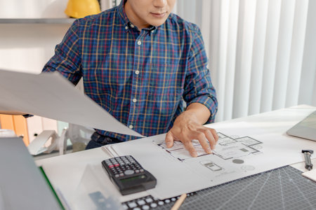 A young architect is working at his desk, reviewing blueprints, using a calculator, and checking a house model. He focuses on precision and detail while planning construction projects.の写真素材