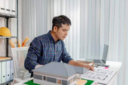A young architect is working at his desk, reviewing blueprints, using a calculator, and checking a house model. He focuses on precision and detail while planning construction projects.の写真素材