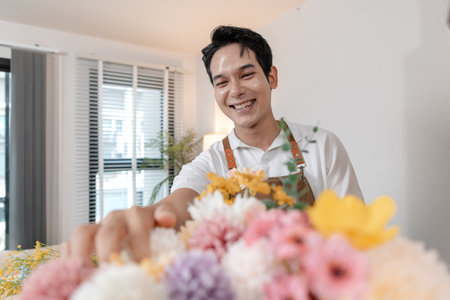 A smiling man wearing an apron arranges colorful flowers in a vase on a table in a cozy room. He appears focused and content while creating a beautiful floral arrangement as part of his work.の写真素材