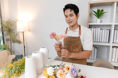 A smiling man wearing an apron arranges colorful flowers in a vase on a table in a cozy room. He appears focused and content while creating a beautiful floral arrangement as part of his work.の写真素材