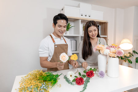 A young couple arranging colorful flowers together in a cozy home setting. The man, wearing an apron, helps the woman create a beautiful bouquet, sharing smiles and creativity in harmony.の写真素材