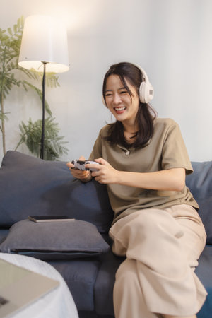 A young woman enjoys playing video games at home, wearing headphones and holding a controller with excitement. She sits comfortably on a sofa, smiling and immersed in digital entertainment.の写真素材