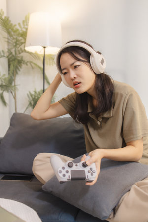 A young woman wearing headphones looks frustrated while playing video games at home. She holds a...