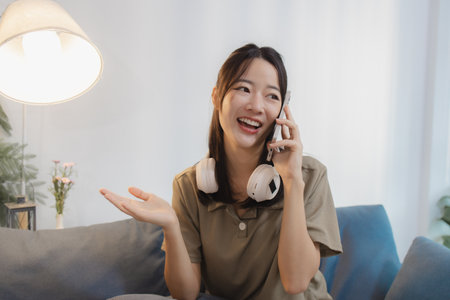 A cheerful young woman sits casually on a sofa, holding a smartphone to her ear while talking and smiling. With headphones around her neck, she looks relaxed, expressive, and engaged.の写真素材