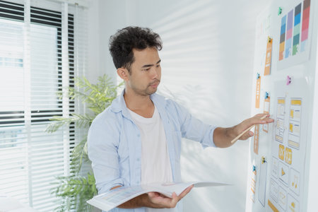 A creative designer is working on UX UI planning, reviewing wireframes, and adjusting color schemes for a mobile application project, showing focus and innovation in modern workspace.の写真素材