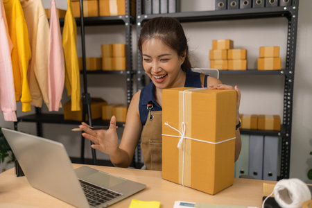 Young female entrepreneur smiling while holding a parcel and using a laptop at her home office. She...