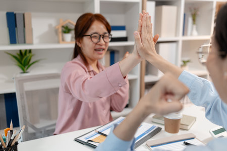 Two Asian businesswomen celebrating success in a modern office after achieving their team goal. They are giving each other a high five with smiles, showing teamwork and positive energy at work.の写真素材
