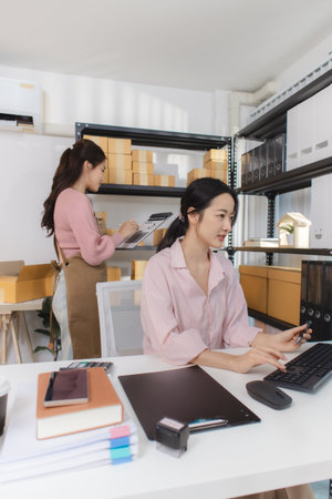 Two young women work together in a small business office, surrounded by shipping boxes and documents. They discuss orders, prepare packages, and manage inventory on a computer.の写真素材