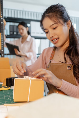 Two young women working together in a small business office, preparing cardboard boxes, checking inventory, and handling packaging tasks as part of their startup online store operations.の写真素材