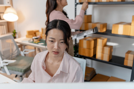 Two young women working together in a small business office, organizing packages, reviewing documents, and planning tasks with smiles. Scene reflects teamwork, productivity and modern entrepreneurshipの写真素材