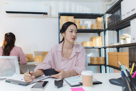 Two young women working together in a small business office, managing online sales and packaging orders. One checks financial reports and uses a calculator, while the other prepares boxes.の写真素材